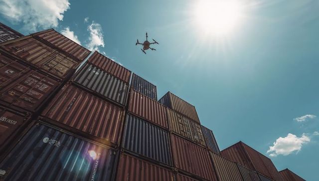 Hovering quadcopter surveying stacked cargo containers under bright sunlight at port yard
