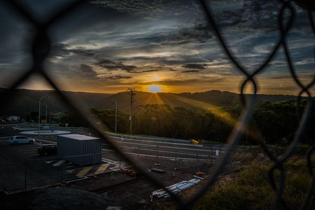 Dramatic sunset behind hills viewed through a wire fence, highlighting a highway and construction area in the foreground. Dusky lighting creates an interesting contrast between the vibrant sky and the darkening surroundings. Ideal for use in projects related to travel, contemplation, nature, and contrast between natural beauty and human infrastructure. Great for metaphoric visuals about barriers, perspective, and resilience.