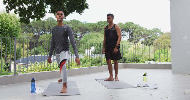 Two young men practicing mindfulness yoga poses outdoors on a terrace with a scenic background. Ideal for illustrating health and wellness concepts, fitness routines, relaxation techniques, or outdoor exercise programs. The image conveys a sense of calm and focus, making it perfect for use in fitness blogs, wellness websites, or as part of a guided yoga program promotion.