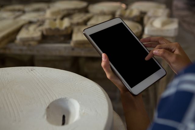 Female potter using digital tablet in a pottery studio. Image shows hands interacting with the device, surrounded by ceramic workpieces. Useful for illustrating the integration of technology in traditional crafts, innovation in art, and creative processes.