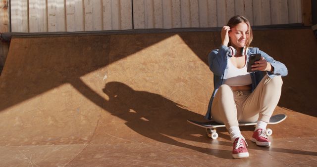 Image of happy caucasian female skateboarder resting and using smartphone. Skateboarding, sport, active lifestyle and hobby concept.