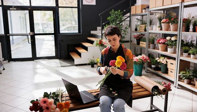 Florist arranging vibrant bouquet on bench trimming stems with shears laptop in bright flower shop