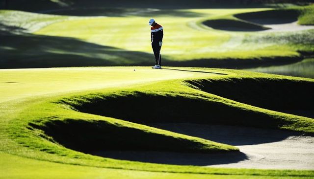 Male golfer lining up putt on sunlit green with sculpted bunkers and rolling fairways