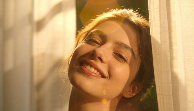 Sunlit woman smiling with eyes closed leaning by sheer curtains during golden hour warmth