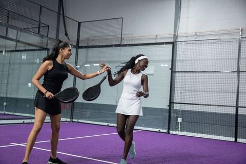 Smiling black and south asian women celebrating on purple padel court in athletic dresses