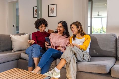 Three friends sharing smartphone and drinks on gray sectional sofa laughing and relaxing