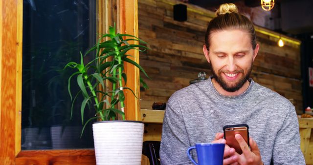 Bearded man with blond hair and man bun seated casually in a cozy café, smiling while looking at his phone. A coffee cup and plant are in the foreground. Ideal for advertising life-style products, mobile technology, or coffee shop ambiance.