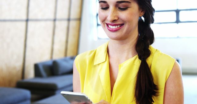 Woman in a bright yellow dress is typing on her smartphone while smiling in a stylish, modern living room. Ideal for illustrating concepts of technology use, modern communication, casual lifestyle, or the joy of staying connected in contemporary living spaces.