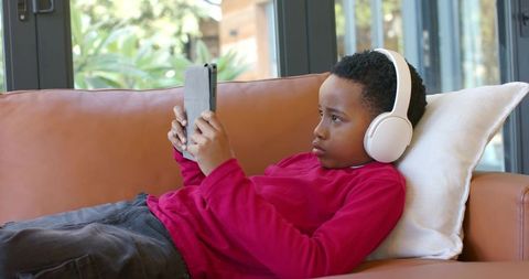 African american boy relaxing on sofa with headphones using tablet in bright living room