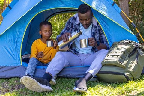 African american father and son pouring hot drink at tent entrance during camping morning