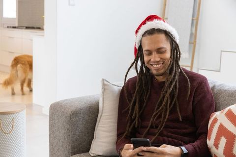 African american man wearing santa hat relaxing on sofa checking smartphone for holiday