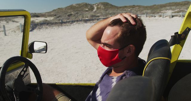 Man sitting in an off-road vehicle wearing a red face mask at beach. Good for travel blogs, pandemic-related content, and summer adventure promotions. Ideal for advertising masks, beach gear, and vacation packages.