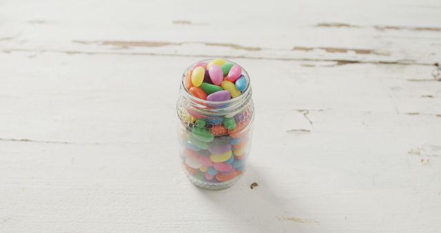 Colorful candy fills a glass mason jar resting on a rustic white wooden table. The vibrant colors of the candy contrast beautifully with the weathered surface, creating a visually appealing scene. Useful for advertisements, blogs, or websites related to sweets, desserts, or rustic aesthetics.