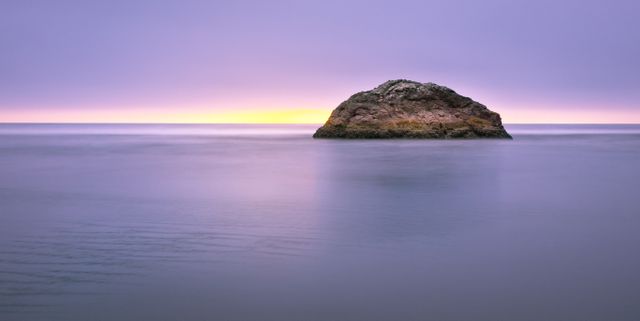 Solitary rocky island in the middle of a calm ocean during twilight with serene waters. Perfect for use in travel promotions, meditation blogs, or nature photography showcases. Highlights themes of serenity, isolation, and the natural beauty of ocean landscapes.