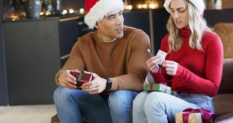 Diverse couple exchanging christmas gifts on couch wearing santa hats, inspecting gift tags
