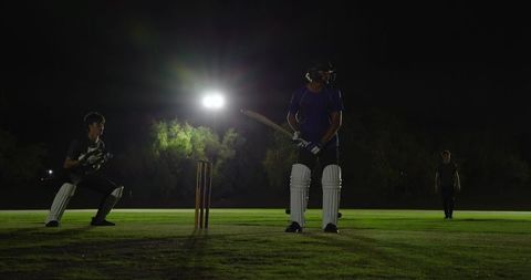 Night cricket batsman standing at crease under floodlight with keeper crouching at stumps