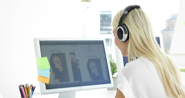 Blonde woman wearing headphones working on computer in creative workspace, possibly engaging in photo editing or graphic design. Usable for office workplace concepts, creativity, technology, business environments, and concentration.