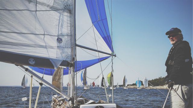 An experienced sailor is navigating through the open sea during a sailing competition with other yachts seen in the background under a clear, sunny sky. Ideal for use in content related to maritime activities, sailing events, nautical training, or adventure and outdoor sports.