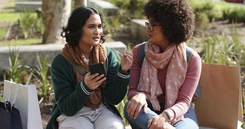 Student friends chatting and sharing smartphone on sunlit campus bench with shopping bags