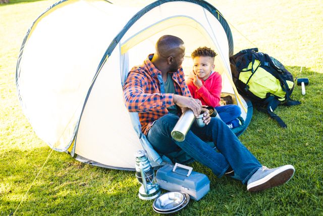 Happy african american father and son sitting in tent and drinking from thermos in their backyard - Download Free Stock Photos Pikwizard.com