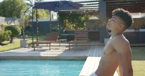 Sunlit african american man relaxing on pool deck by backyard pergola and lounge chairs