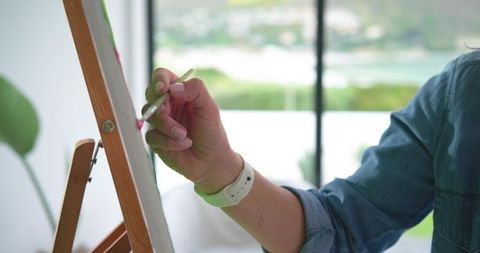 Female artist painting on canvas at easel closeup of hand holding brush in sunlit studio