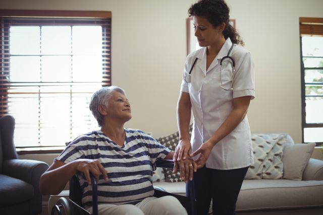 Female doctor providing assistance to a senior woman in a wheelchair at home. Ideal for use in healthcare, elderly care, home care services, and medical support content. Highlights compassionate care and professional medical assistance in a home setting.