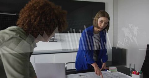 Asian woman pointing at blueprints and reviewing plans with colleague in modern boardroom