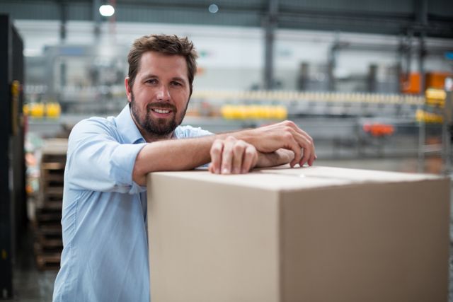 Smiling factory worker leaning on a large cardboard box in an industrial warehouse. Ideal for use in articles or advertisements related to manufacturing, logistics, labor, and workplace environments. Can be used to depict themes of hard work, job satisfaction, and industrial operations.