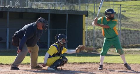 Sunlit baseball batter preparing to swing with catcher and umpire at home plate during amateur game