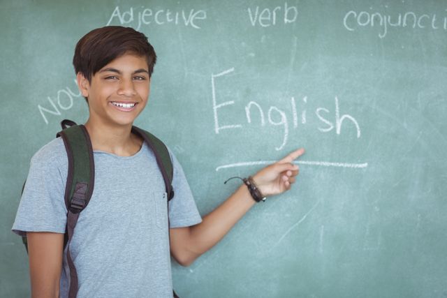 Schoolboy standing in classroom pointing at chalkboard with English written on it. Ideal for educational materials, school promotions, academic websites, and learning resources.