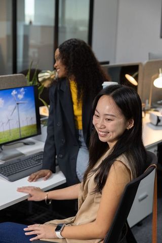 Smiling diverse female coworkers collaborating in modern office with wind turbine display on monitor