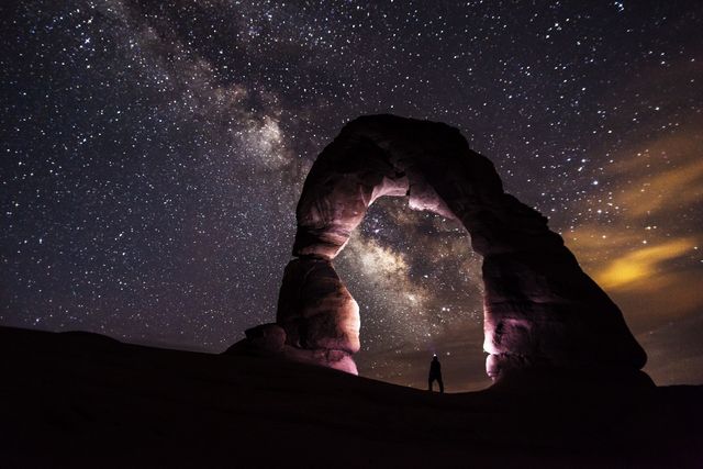 Silhouette of a person standing beneath Delicate Arch with Milky Way stretching across night sky. Captures beauty of natural arch formations against backdrop of a mesmerizing starlit sky. Ideal for use in travel blogs, astronomy articles, or educational materials about national parks and astro-photography. Perfect for promoting destinations, inspiring wanderlust, or enhancing science communications.