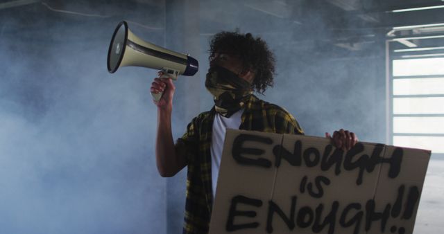 Protester holding megaphone and sign in smoky environment. Sign reads 'Enough is Enough'. Suitable for advocacy, social movements, protest-related content, campaigns against injustice.