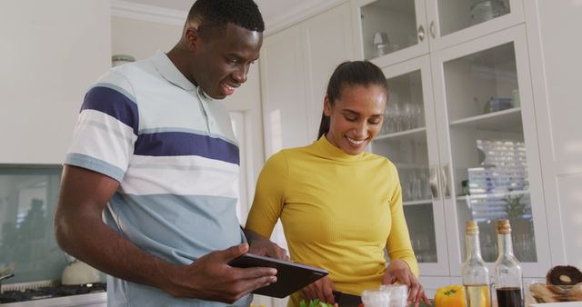Image of happy diverse couple using recipe on tablet preparing food together in kitchen. Communication, happiness, health, domestic life and inclusivity concept.