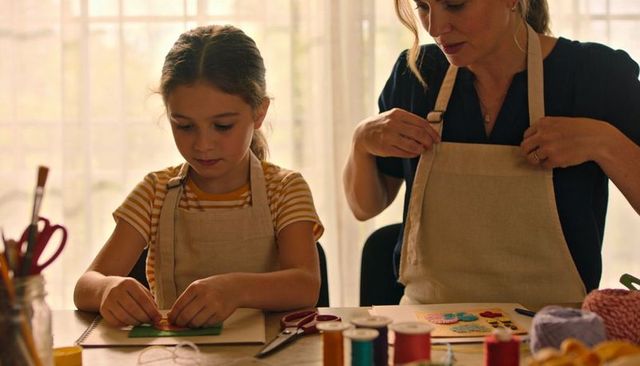 Mother adjusting apron while daughter creating paper craft at sunlit table