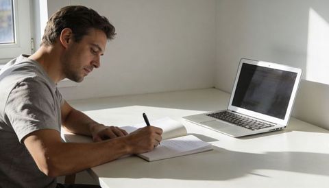 Focused young man writing at minimalist home desk with laptop and notebook in daylight