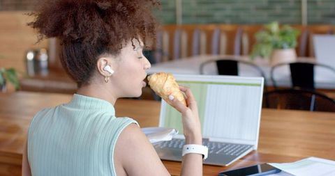 Remote worker eating croissant at cafe with laptop, earbuds and smartwatch on wooden table