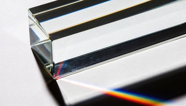 Rectangular glass prism refracting narrow rainbow beam across white background with sharp facets
