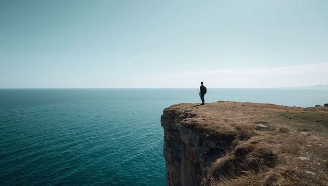 Hiker standing on windswept cliff overlooking calm turquoise ocean horizon
