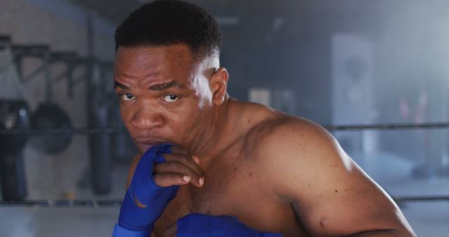 A male boxer with intense focus, standing in a gym holding a punching stance. Dressed in gloves, the setting suggests dedication to training and physical fitness. Perfect for use in content related to sports motivation, fitness coaching, athletic training programs, and boxing promotions.