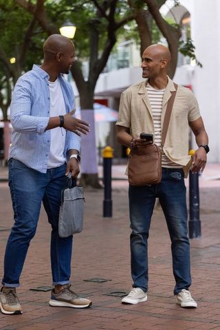 Two african american men walking and talking on urban sidewalk carrying bags and smartphone
