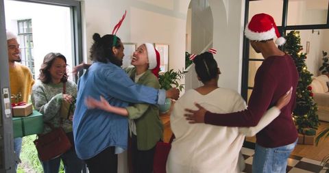Diverse friends greeting with hugs and gifts at festive home entryway wearing santa hats