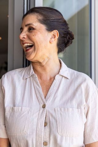Mature woman laughing while turning head left at sunlit sliding glass door in beige shirt