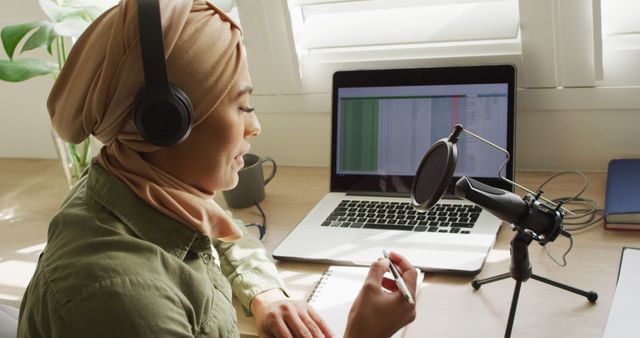 Woman wearing hijab recording podcast at home office desk, using laptop and professional microphone setup while taking notes. This is ideal for content relating to remote work, podcasting, technology in creative industries, home office setups, and professional women. Useful for illustrating multicultural, modern work environments and creative workspaces.