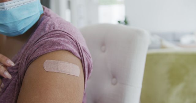 This close-up of an arm with an adhesive bandage after a vaccination is perfect for illustrating health and safety, medical campaigns, or Covid-19 awareness. The individual's use of a face mask adds to the focus on health precautions.
