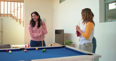 Diverse women playing pool at home laughing around blue-felt billiard table