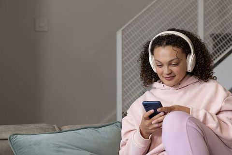 Young woman relaxing on sofa listening to music with headphones while using smartphone