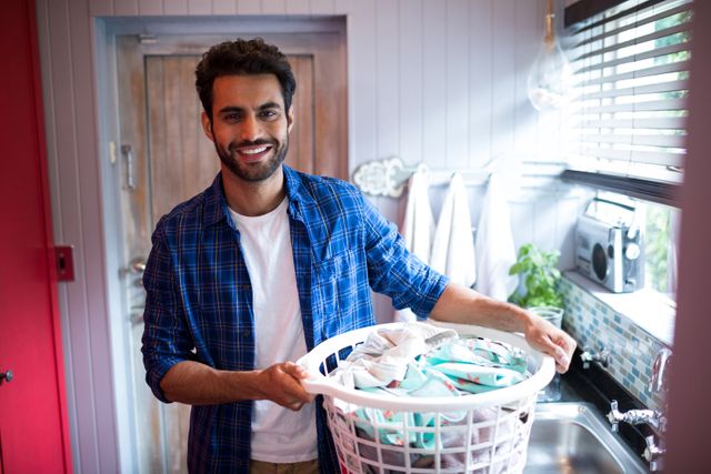 Young man standing by window in bright kitchen, holding laundry basket filled with clothes. Useful for content related to home chores, laundry routines, and domestic lifestyle.
