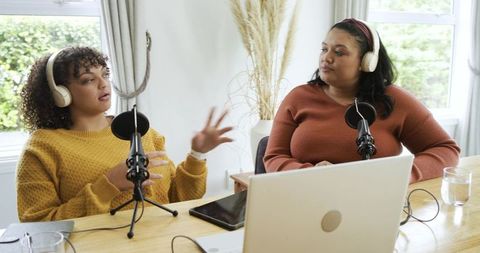 Women podcast hosts wearing headphones recording candid conversation at home table